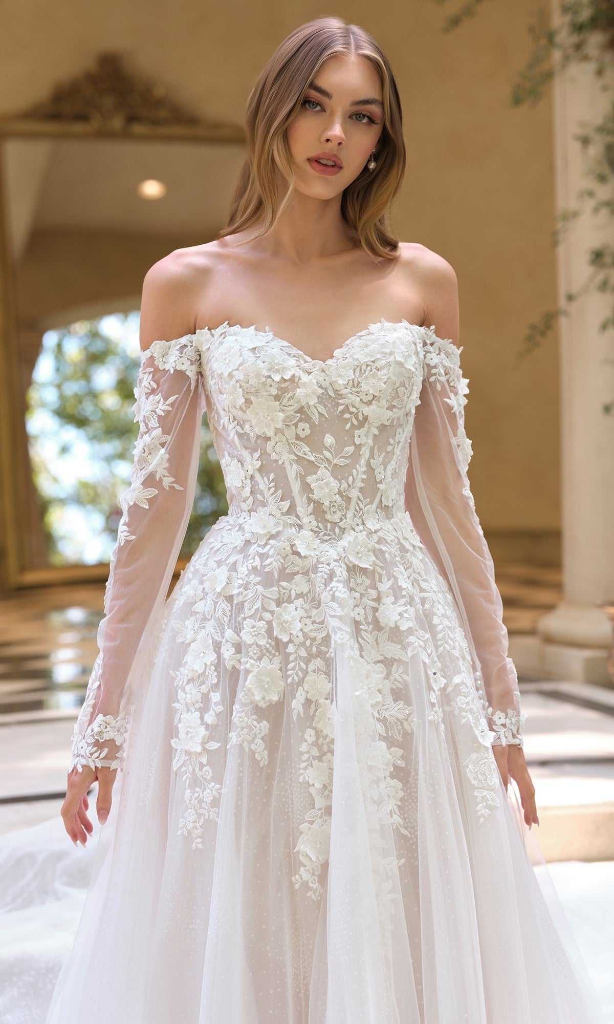 A woman wearing the White Lace Wedding Dress: Andrea and Leo WL067, featuring a corset bodice, long sleeves, and 3D floral lace applique, stands indoors near a large window.